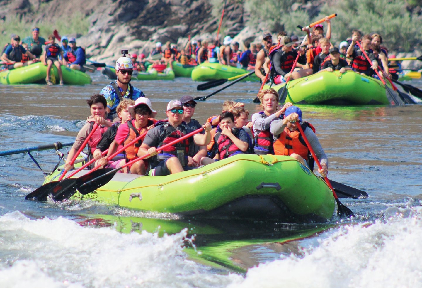 A loaded paddle raft driving through a rapid on the Salmon River with a full crew digging in, several more rafts working the same water behind it and a crowd of guests waiting along the bank.