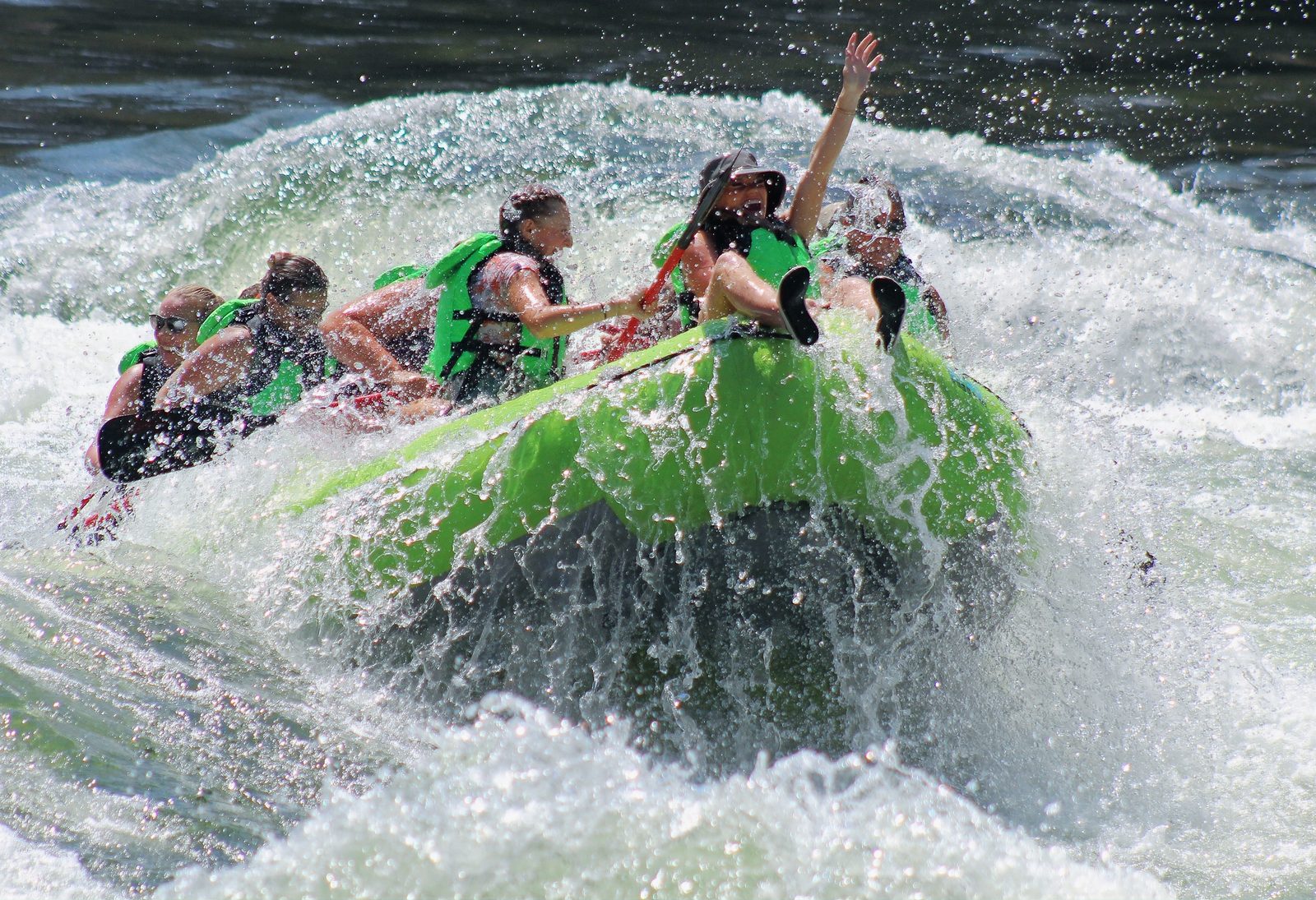 A green raft full of guests punching through a large whitewater wave on the Salmon River, one passenger with an arm raised.