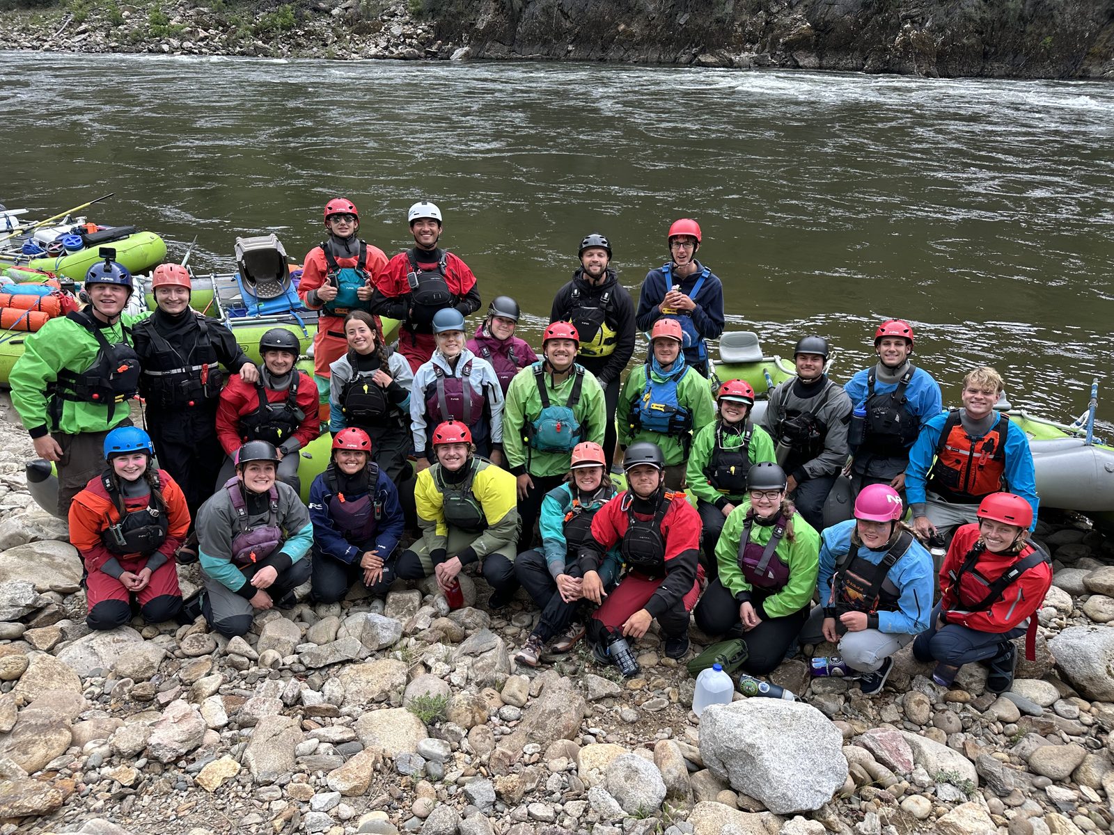 Roughly thirty Adventure Idaho river guides in drysuits and helmets gathered on a rocky beach beside rafts on the Salmon River in Idaho.