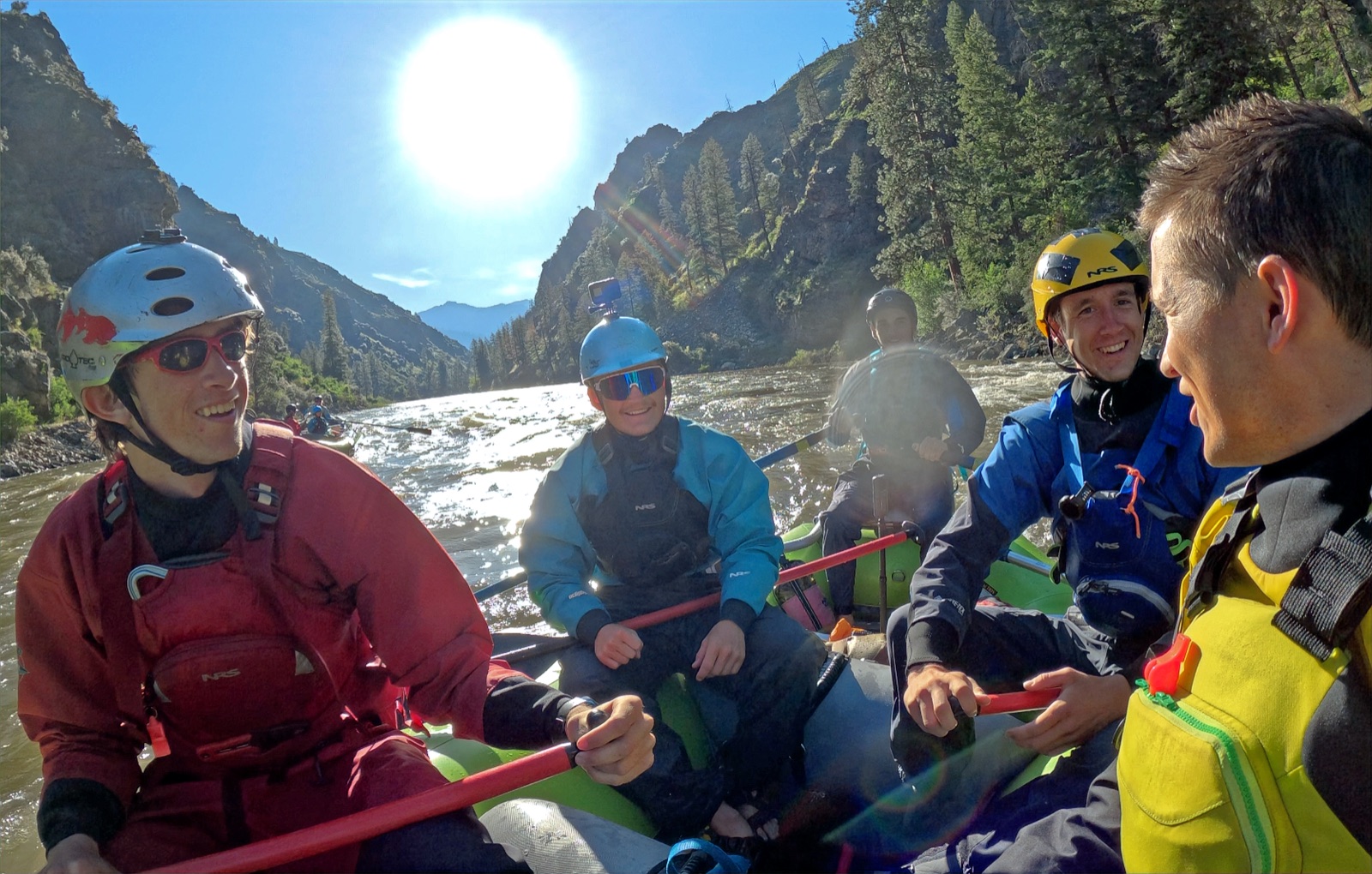 Justin Smith and four Adventure Idaho guides in drysuits and helmets sitting in a raft on the Salmon River in morning light, talking through a training run.